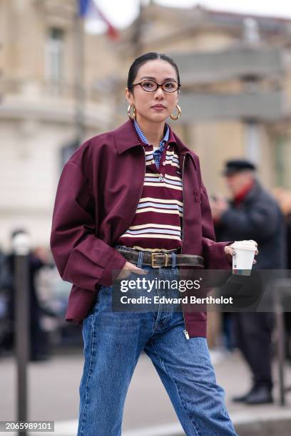 Yoyo Cao wears golden earrings, a burgundy oversized jacket , a striped polo shirt, a belt, blue denim pants / jeans , outside Miu Miu , during the...