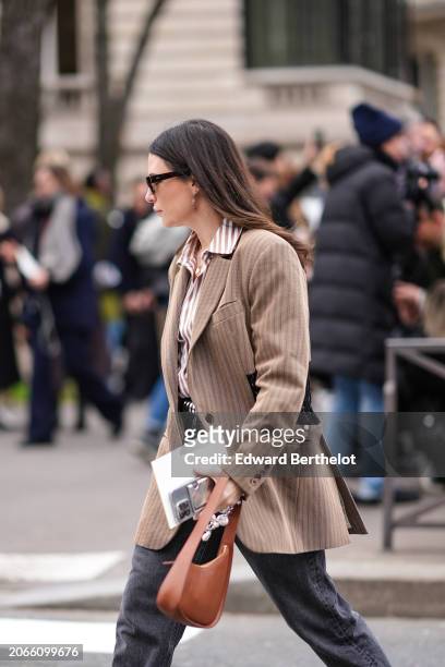Guest wears sunglasses, earrings, a beige striped oversized blazer jacket, a striped shirt, a brown leather bag , outside Miu Miu , during the...