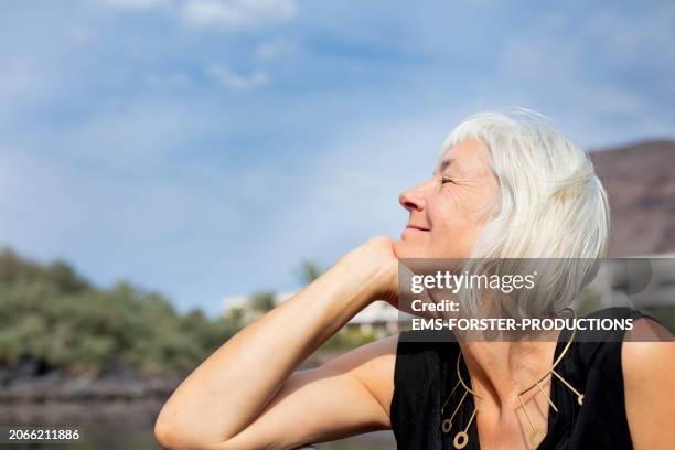 portrait of a beautiful senior woman sitting on beach and enjoying the sun in la gomera - white hair stock-fotos und bilder