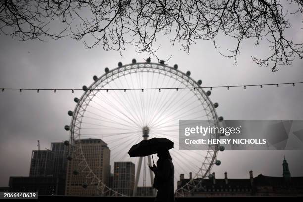 Pedestrian shelters from the rain beneath an umbrella, backdropped by the landmark the London Eye, in London on March 10, 2024.