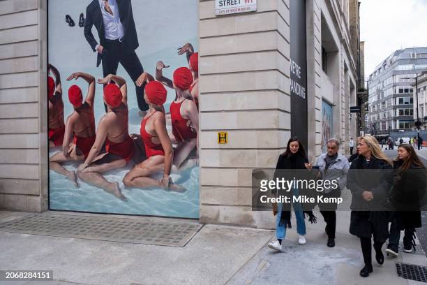 Advertising imagery featuring several women in formation wearing red bathing suits and swimming caps looking up to a male wearing a suit for the...