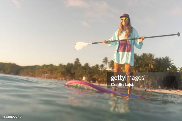 a smiling woman keeps her balance while standing on a stand-up paddle board - paddling stock pictures, royalty-free photos & images