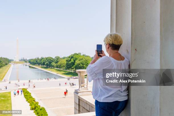 a mature blonde woman photographing the washington monument and reflecting pool from the lincoln memorial entrance in washington, d.c. - washington monument dc stock pictures, royalty-free photos & images