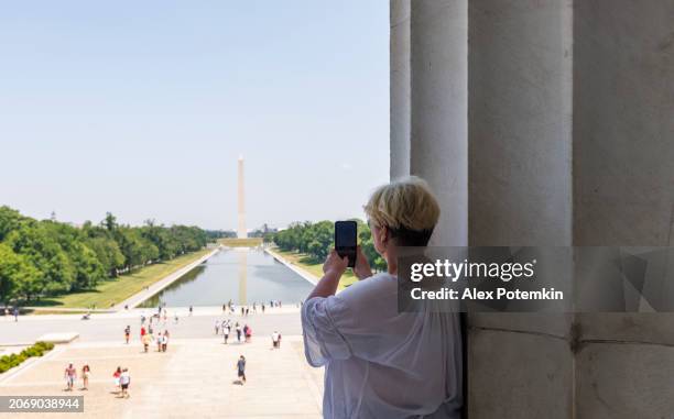 mature woman photographing washington monument with reflecting pool from lincoln memorial on a sunny summer day in washington, d.c. - washington monument dc stock pictures, royalty-free photos & images