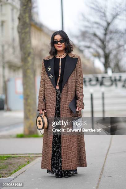 Guest wears sunglasses, a necklace, a black cardigan, a brown long coat, a Chanel bag, flared pants in black and white with floral print , outside...