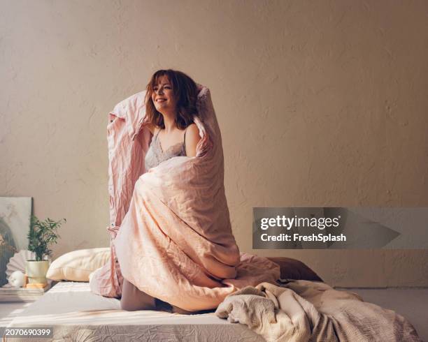 smiling woman wrapped in comforter enjoying cozy morning at home - dekbed stockfoto's en -beelden