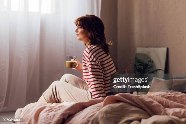 donna contemplativa che si gode il caffè mattutino nella camera da letto illuminata dal sole - lentezza foto e immagini stock