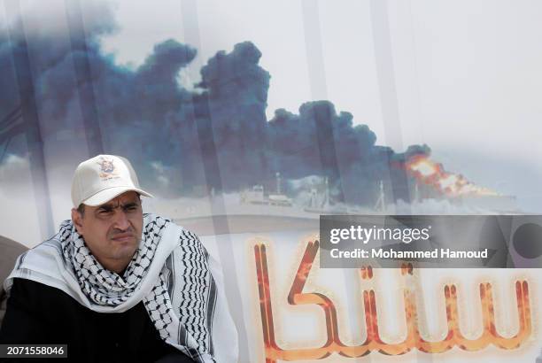 Yemen's Houthi follower sits next to a billboard bearing the image of a burning commercial ship targeted previously by Houthi forces in the Red Sea...