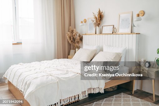 Simple bulb lamp on a rope hanging above bed with white bedclothes, books and gold fern leaf on an end table in white bedroom interior Simple bulb lamp on a rope hanging above bed with white bedclothes, books and gold fern leaf on an end table in white bedroom interior