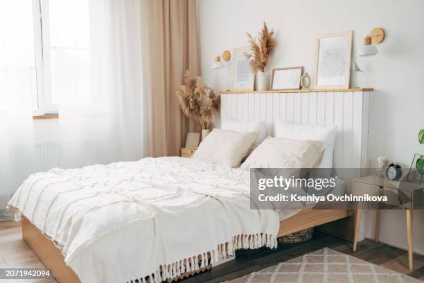 simple bulb lamp on a rope hanging above bed with white bedclothes, books and gold fern leaf on an end table in white bedroom interior - kussen beddengoed stockfoto's en -beelden