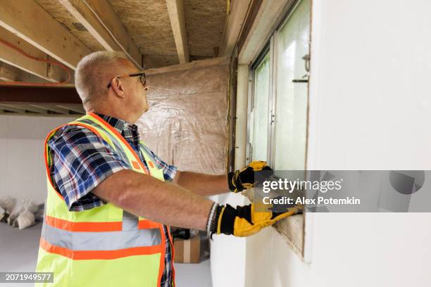 el agente de reclamos revisa las ventanas del sótano para verificar si son resistentes. inspector de seguros maduro con chaleco de seguridad, guantes y anteojos midiendo y revisando las ventanas del sótano - flooded basement fotografías e imágenes de stock