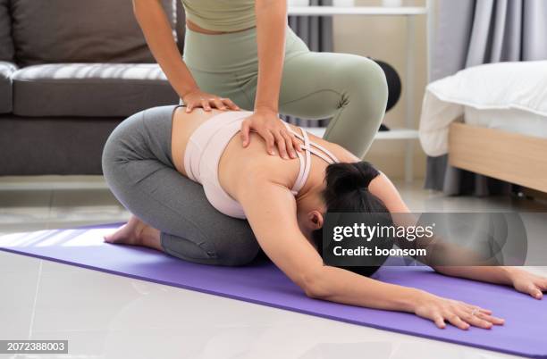 Two Asian women practicing yoga exercise with partner in bedroom at home. Trainer helps student to do Balasana exercise or Child pose