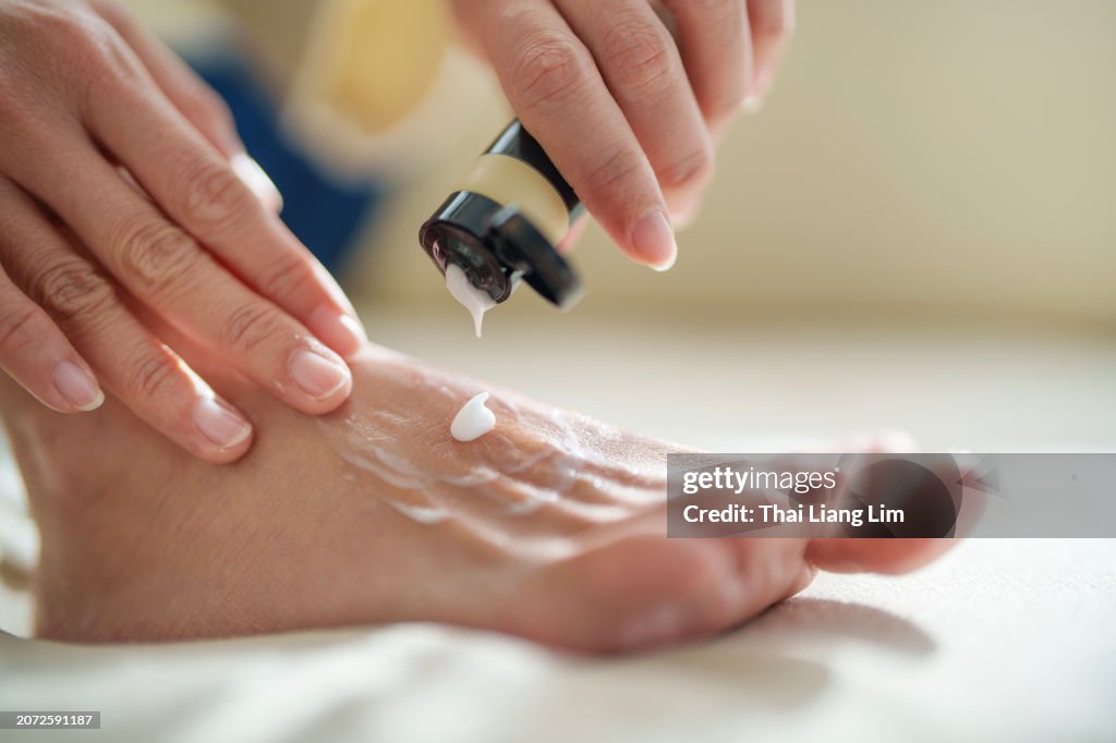 In the cropped image, a mid-adult Asian woman is shown applying moisturizing cream to her dry skin on her feet, while the sunlight gently shines on her skin, highlighting the nurturing gesture.