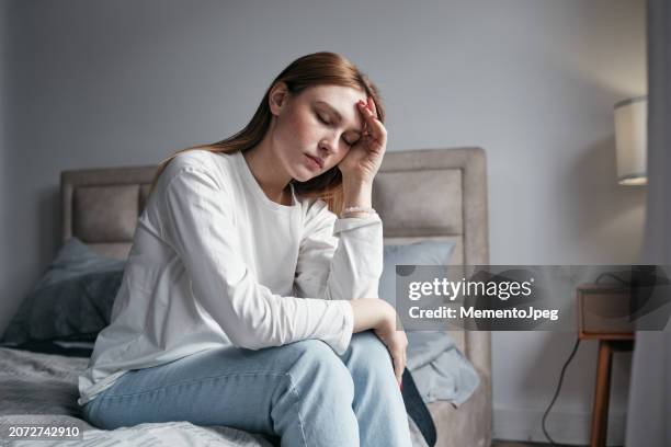 morning headache. young woman sitting on bed with eyes closed touching head suffering from headache or migraine, feeling exhaustion - burnout stockfoto's en -beelden