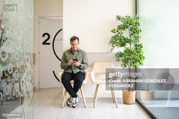 man using mobile sitting in a waiting room of a clinic - hospital waiting room stockfoto's en -beelden