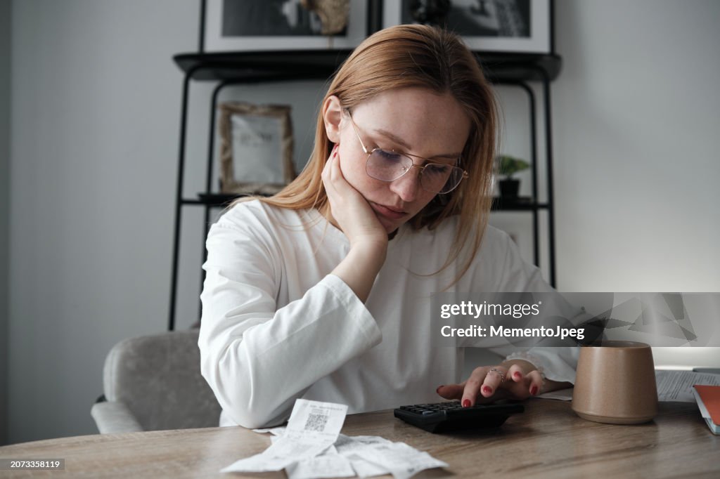 Young woman calculating spendings looking at bills using calculator while sitting at table. Cost of Living