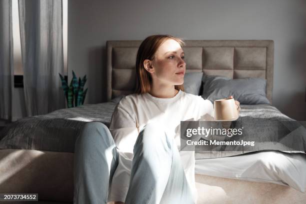 slow living. happy pleased young woman with cup of morning coffee, enjoying sunlight trough windows. redhead millennial girl taking break resting at home drinking tea - slow living photos et images de collection