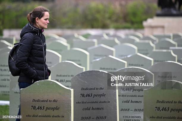 Woman walks among gravestones reading "Dedicated to the 70 463 people who died avoidable death in cold homes 2013-2023, cold homes cost lives" and...