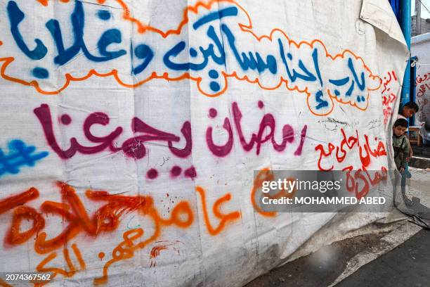 Palestinian boys stand near a tarp bearing a greeting message for the Muslim holy month of Ramadan beneath another prayer pleading God in Arabic, at...