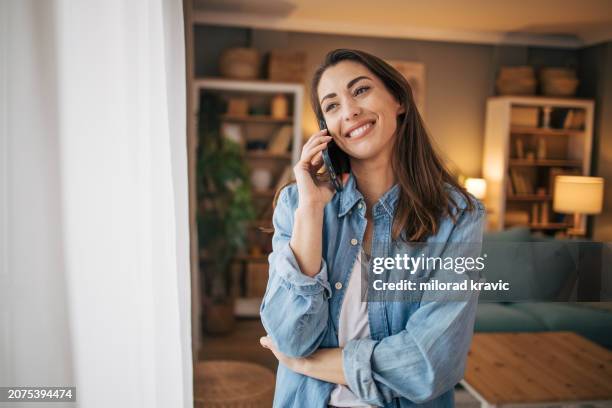 foto einer frau, die mit einer freundin telefoniert, während sie im wohnzimmer steht - am telefon stock-fotos und bilder