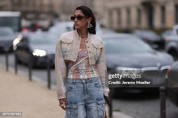 Guest seen wearing brown Chanel sunglasses, triangular and silver shimmery Chanel earrings, a tweed creme jacket, a colorful shirt, denim blue jeans,...