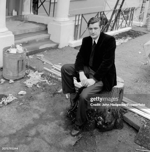 English poet and writer Ted Hughes seated on a tree stump on a residential street in London in 1959.