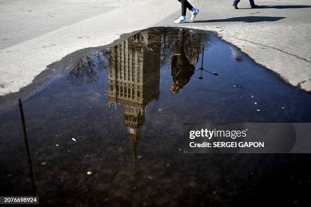 Woman is reflected in a puddle as she walks in front of the Palace of Culture and Science during a sunny day in Warsaw, Poland on March 14, 2024.