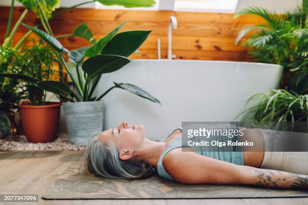 post-meditation repose: grizzled natural woman lies in corpse pose, relaxing after yoga in bright green bathroom - bad breath stockfoto's en -beelden