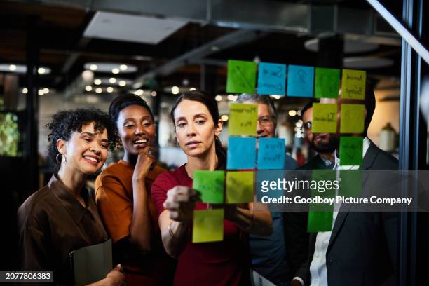 lächelnde geschäftsleute brainstormen mit klebenotizen an einer glaswand in einem büro - brainstormen stock-fotos und bilder