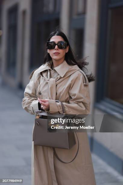 Tugba Kement seen wearing YSL black sunglasses, COS beige oversized long coat, Hermès Kelly taupe / brown leather bag and gold rings, on March 08,...