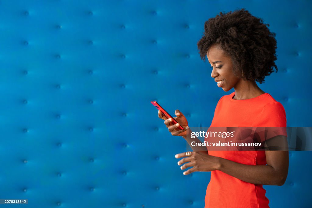 Studio shot of an attractive african young woman, wearing red t-shirt, posing in front of blue background and talking on a smart phone