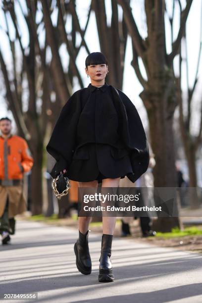 Akimoto Kozue wears a black oversize gathered jacket worn as a mini dress, black leather boots, a black bag with golden chains, outside Sacai, during...