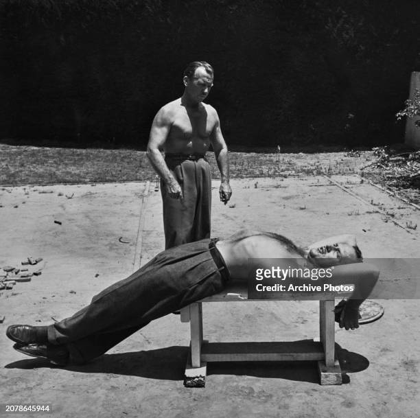 American actor Keenan Wynn, shirtless and lying on a weight bench, lifting weight as a second shirtless man stands over him, during production of the...