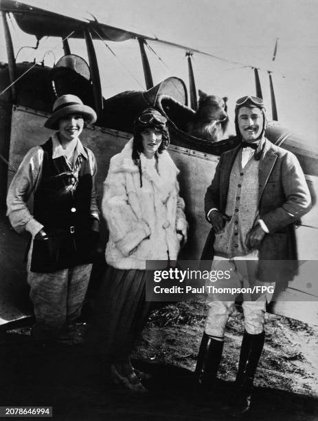 American stunt pilot and actor Al Wilson and his wife, Ruth Mitchell, pose in aviator's caps and goggles beside an aircraft, a dog sits in the...