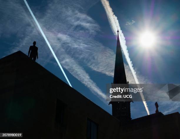 Anthony Gormley public sculpture on a roof in Oxford, dramatic sky with sun and contrails.