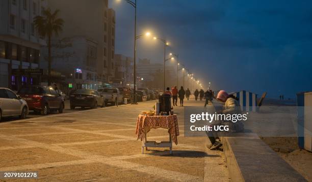 Tunisian man sits on the promenade and looks at his smartphone as he waits to sell tea to passers by next to Bou Jaafar beach in Sousse, Tunisia.