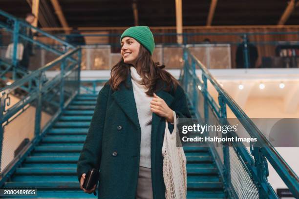 low angle shot of beautiful woman walking down the stairs, holding ecological shopping bag with groceries. woman in the city shopping at thrift market. concept of recycling, upcycling and environmental sustainability. - woolly hat stock pictures, royalty-free photos & images