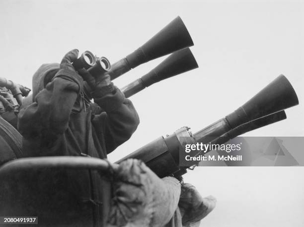 Royal Navy sailor scans the sky with binoculars searching for German Luftwaffe aircraft beside a quad mount Vickers QF 2-pounder MkVII pom-pom...