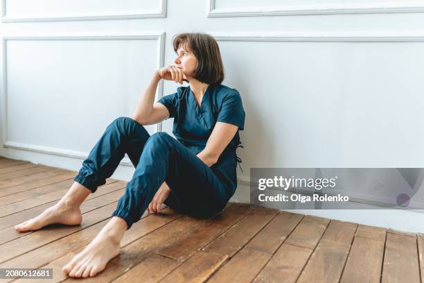 female doctor in a blue medical uniform sits near the wall on the floor and looks out the window. - flooring stock pictures, royalty-free photos & images