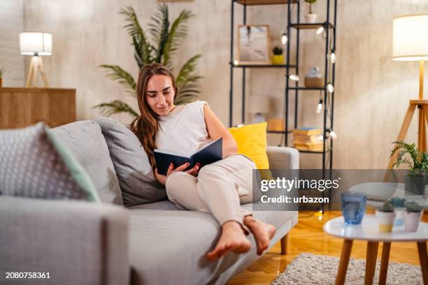 young woman sitting on the sofa and reading a book - escaping room stock pictures, royalty-free photos & images