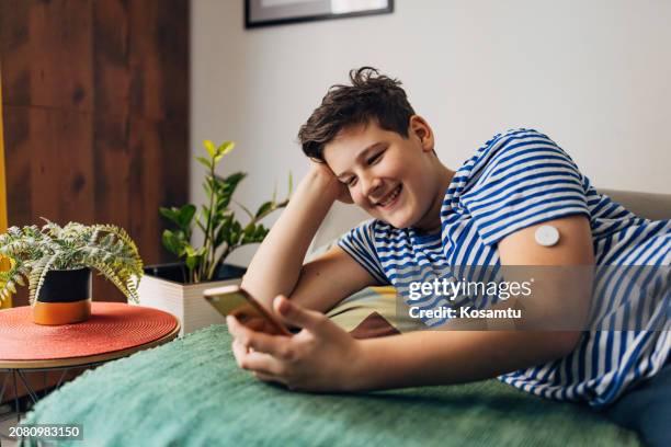 a cheerful boy with type 1 diabetes uses a mobile phone to surf the internet while relaxing at home - continuous glucose monitor stock pictures, royalty-free photos & images