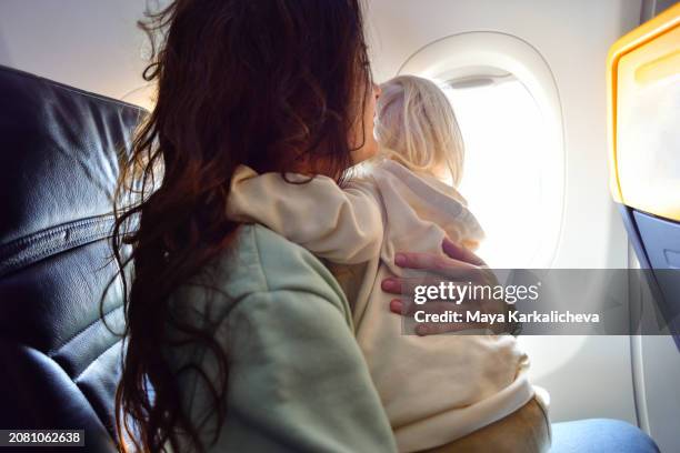 mother embracing her baby while looking through window on airplane - woman blond looking left window stockfoto's en -beelden