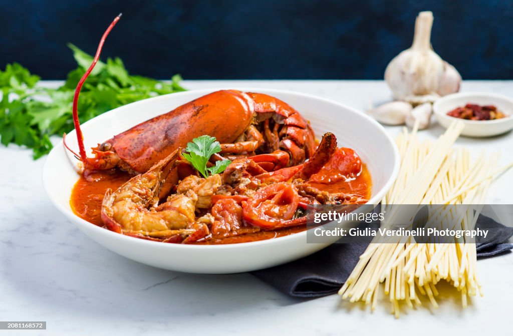 Bowl of lobster stew sauce with whole lobsters and tomato sauce on a white marble table with ingredients in the background