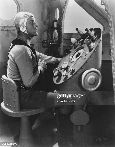American swimmer and actor Buster Crabbe in costume as he sits at a control panel in a scene from 'Flash Gordon', filmed at Universal Studios in...