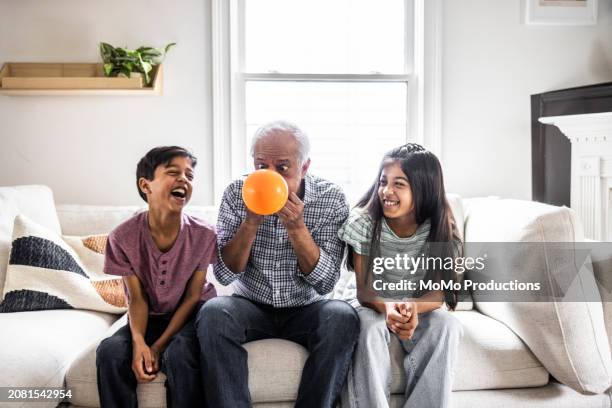 grandfather blowing up balloon while grandchildren laugh - poniendo muecas de broma fotografías e imágenes de stock