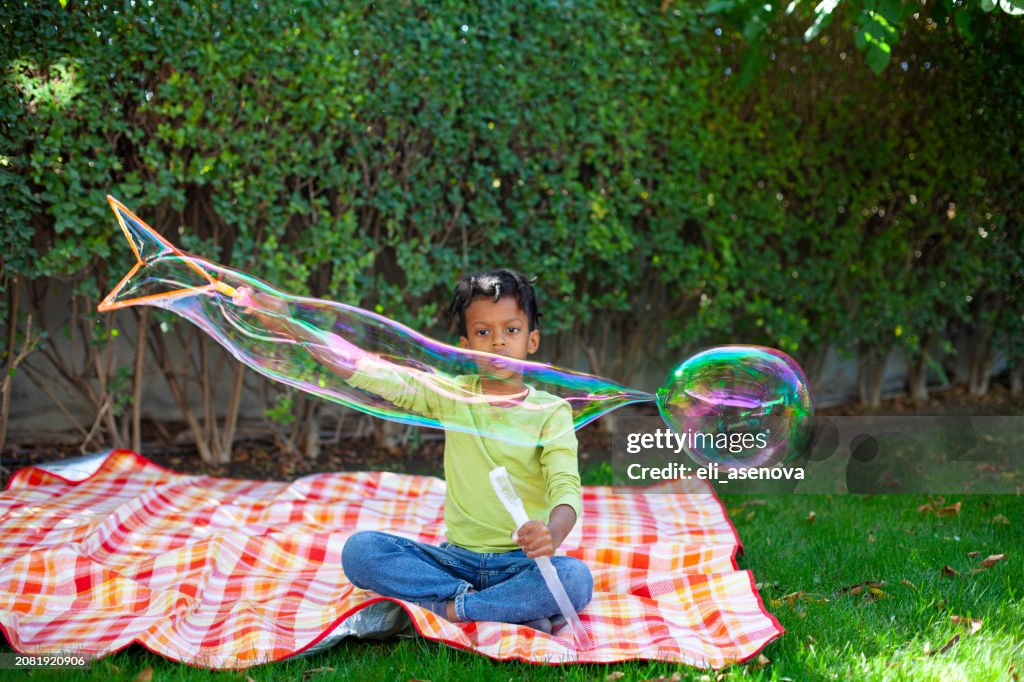 Happy African-American boy with black curly hair blowing and playing with soap bubbles at green garden
