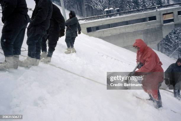 Man wearing waterproof clothing sprays water on snow as soldiers prepare the St Nizier ski jump slope for the 1968 Winter Olympics, near Grenoble,...