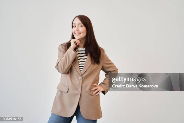 a business woman in a beige suit and jeans stands on a monochromatic background smiles and looks at camera inspiredly with copy space. - beige jacket stock pictures, royalty-free photos & images