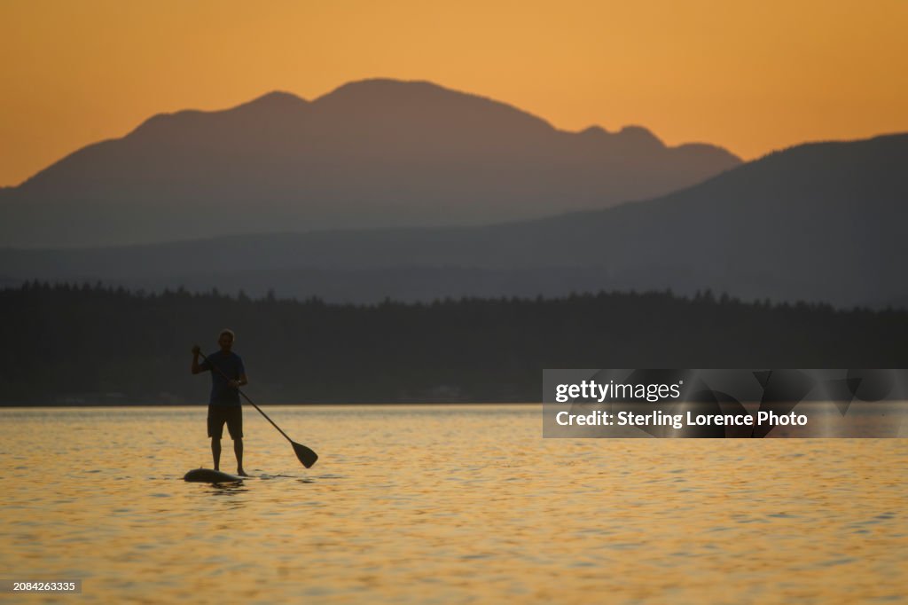 A senior citizen man goes for an evening work out paddleboarding and to watch a dramatic sunset near his recreational property in the gulf islands of British Columbia near Gabriola Island, Valdez island, Vancouver Island, Nanaimo. warm summer holiday