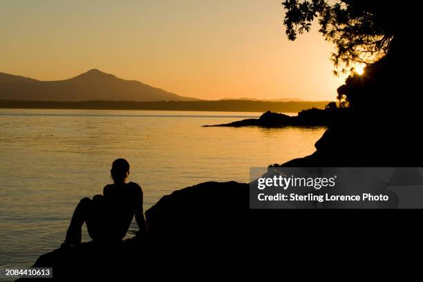 a woman sits on a sandstone bluff in the gulf islands of british columbia to watch a summer sunset over the pacific ocean and the distant mountains of vancouver island near nanaimo, salt spring island, ruxton island, gabriola island and thetis island. - gabriola isle stockfoto's en -beelden